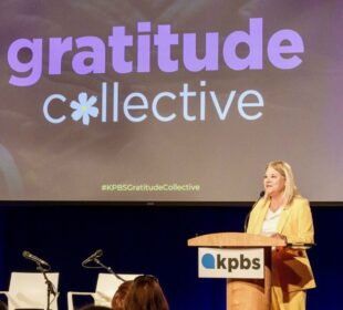 Wearing a yellow suit, Deanna Mackey, GM of KPBS in San Diego, speaks at a podium with the KPBS logo in front of a large screen displaying the words “gratitude collective” with a flower symbol replacing the “o.” The event is a formal gathering, with an audience seated and additional microphones and chairs on stage.