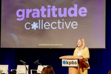 Wearing a yellow suit, Deanna Mackey, GM of KPBS in San Diego, speaks at a podium with the KPBS logo in front of a large screen displaying the words “gratitude collective” with a flower symbol replacing the “o.” The event is a formal gathering, with an audience seated and additional microphones and chairs on stage.