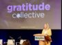 Wearing a yellow suit, Deanna Mackey, GM of KPBS in San Diego, speaks at a podium with the KPBS logo in front of a large screen displaying the words “gratitude collective” with a flower symbol replacing the “o.” The event is a formal gathering, with an audience seated and additional microphones and chairs on stage.