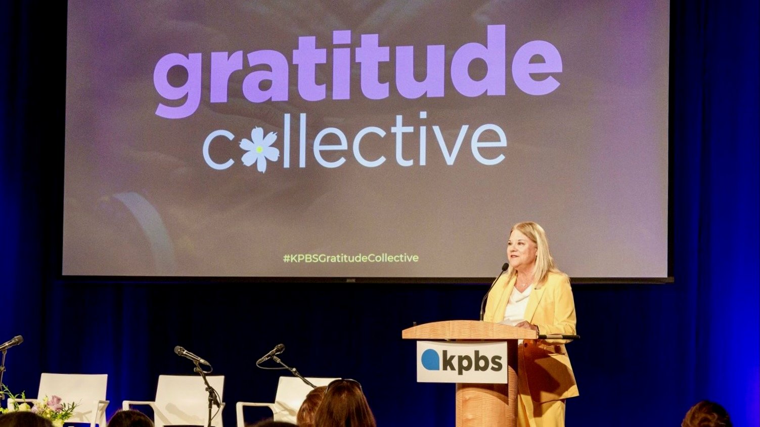 Wearing a yellow suit, Deanna Mackey, GM of KPBS in San Diego, speaks at a podium with the KPBS logo in front of a large screen displaying the words “gratitude collective” with a flower symbol replacing the “o.” The event is a formal gathering, with an audience seated and additional microphones and chairs on stage.