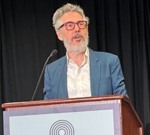 Ira Glass stands at a podium speaking into a microphone, wearing a blue blazer, white shirt, and red eyeglasses. A black curtain hangs in the background, and a circular logo is partially visible on the front of the podium.