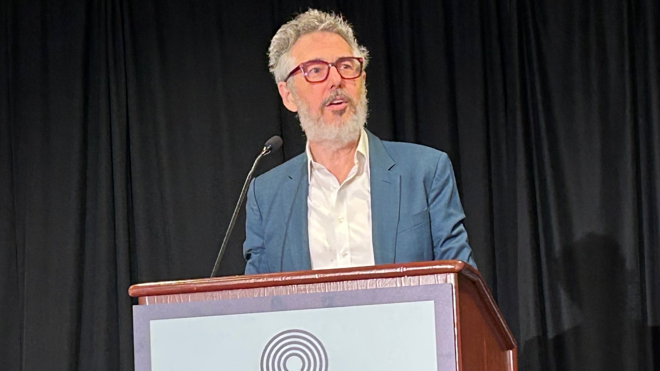 Ira Glass stands at a podium speaking into a microphone, wearing a blue blazer, white shirt, and red eyeglasses. A black curtain hangs in the background, and a circular logo is partially visible on the front of the podium.