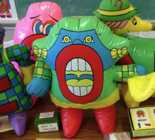 Colorful inflatable Letter People characters are displayed on a classroom table alongside reading materials and board games. The inflatables represent letters including E, O, and N, each decorated with cartoon faces and costumes.