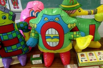 Colorful inflatable Letter People characters are displayed on a classroom table alongside reading materials and board games. The inflatables represent letters including E, O, and N, each decorated with cartoon faces and costumes.