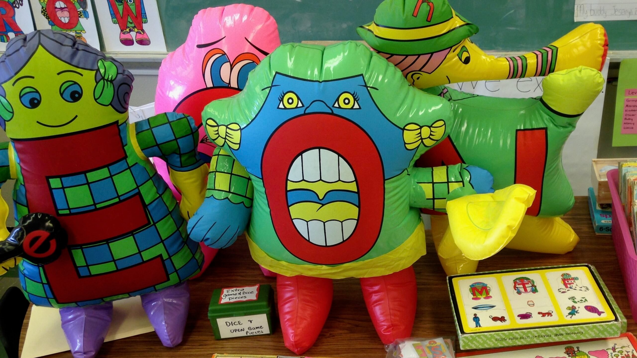 Colorful inflatable Letter People characters are displayed on a classroom table alongside reading materials and board games. The inflatables represent letters including E, O, and N, each decorated with cartoon faces and costumes.
