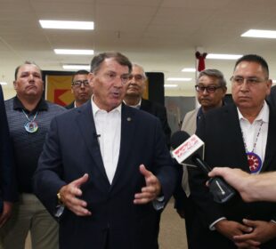 Senator Mike Rounds speaks to reporters while standing in front of a group of Native American leaders. He gestures with his hands as microphones are held toward him. The men behind him wear suits and traditional tribal medallions. The setting appears to be a community or government facility, with overhead lights and visible flags in the background.