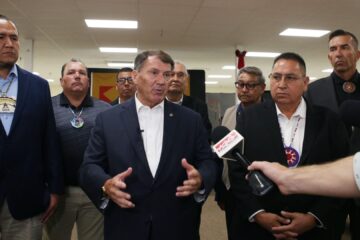 Senator Mike Rounds speaks to reporters while standing in front of a group of Native American leaders. He gestures with his hands as microphones are held toward him. The men behind him wear suits and traditional tribal medallions. The setting appears to be a community or government facility, with overhead lights and visible flags in the background.