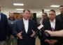 Senator Mike Rounds speaks to reporters while standing in front of a group of Native American leaders. He gestures with his hands as microphones are held toward him. The men behind him wear suits and traditional tribal medallions. The setting appears to be a community or government facility, with overhead lights and visible flags in the background.
