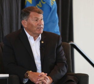 U.S. Sen. Mike Rounds, R-South Dakota, wearing a dark suit and white shirt, sits in a chair speaking during a Greater Sioux Falls Chamber of Commerce event on Aug. 28, 2025. A blue flag and black curtain are in the background, with a banner partially visible at left. Two water glasses sit on the table in front of him.