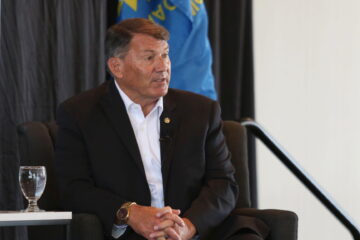 U.S. Sen. Mike Rounds, R-South Dakota, wearing a dark suit and white shirt, sits in a chair speaking during a Greater Sioux Falls Chamber of Commerce event on Aug. 28, 2025. A blue flag and black curtain are in the background, with a banner partially visible at left. Two water glasses sit on the table in front of him.
