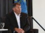 U.S. Sen. Mike Rounds, R-South Dakota, wearing a dark suit and white shirt, sits in a chair speaking during a Greater Sioux Falls Chamber of Commerce event on Aug. 28, 2025. A blue flag and black curtain are in the background, with a banner partially visible at left. Two water glasses sit on the table in front of him.