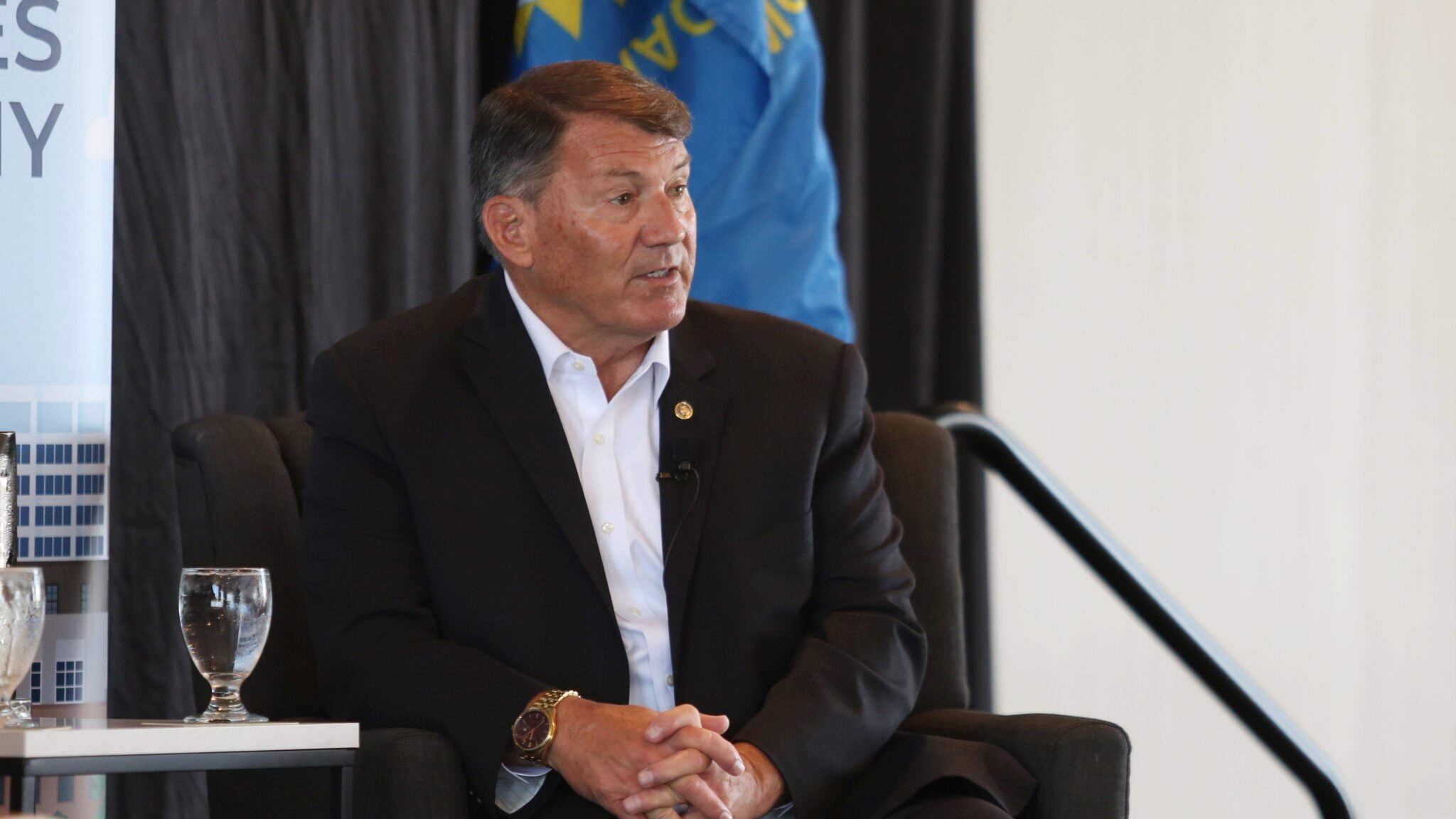U.S. Sen. Mike Rounds, R-South Dakota, wearing a dark suit and white shirt, sits in a chair speaking during a Greater Sioux Falls Chamber of Commerce event on Aug. 28, 2025. A blue flag and black curtain are in the background, with a banner partially visible at left. Two water glasses sit on the table in front of him.