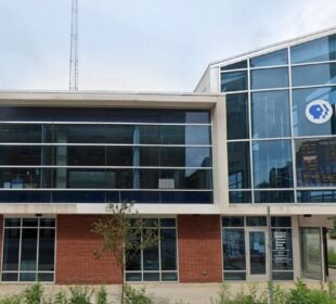 The headquarters of PBS Michiana – WNIT, a modern two-story building with large glass windows and red brick on the ground floor. The PBS logo is displayed prominently on the upper right corner of the building. A handicap parking sign is visible in front, along with a small tree and sidewalk.