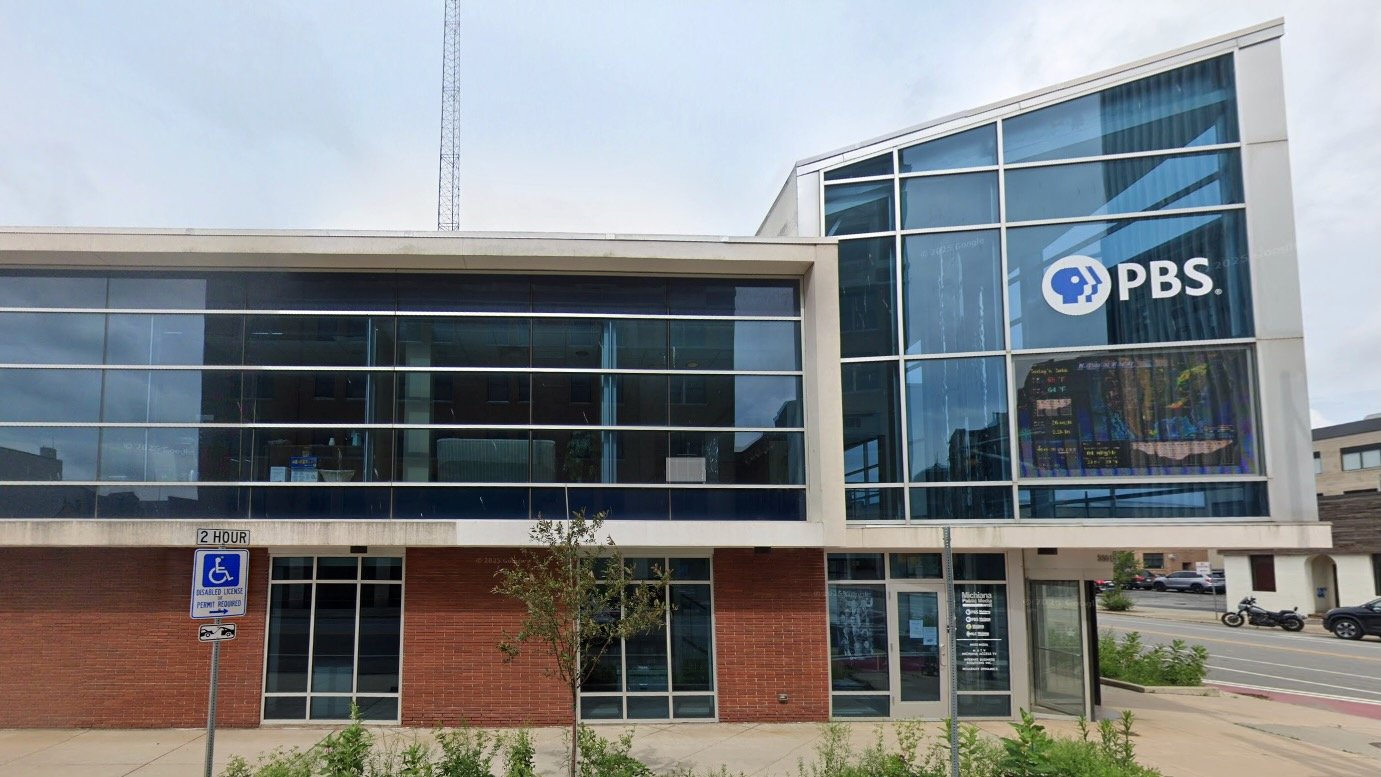 The headquarters of PBS Michiana – WNIT, a modern two-story building with large glass windows and red brick on the ground floor. The PBS logo is displayed prominently on the upper right corner of the building. A handicap parking sign is visible in front, along with a small tree and sidewalk.