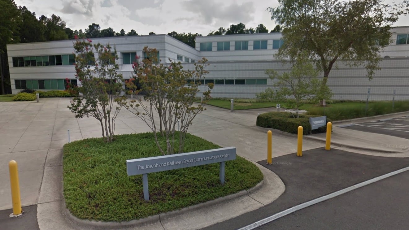 Exterior view of the PBS North Carolina headquarters, known as The Joseph and Kathleen Bryan Communications Center, located in Research Triangle Park. The modern two-story building features a white and gray facade with rows of green-tinted windows, surrounded by trees and landscaped greenery. A sign with the building name is visible in the foreground, near visitor parking.