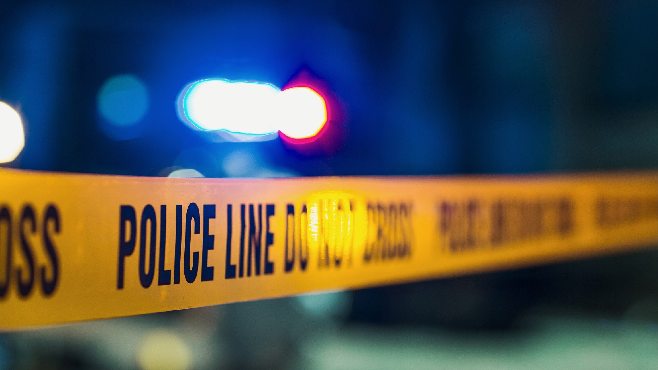 Close-up of yellow police line tape reading “POLICE LINE DO NOT CROSS,” with blurred red and blue emergency lights in the background at night.