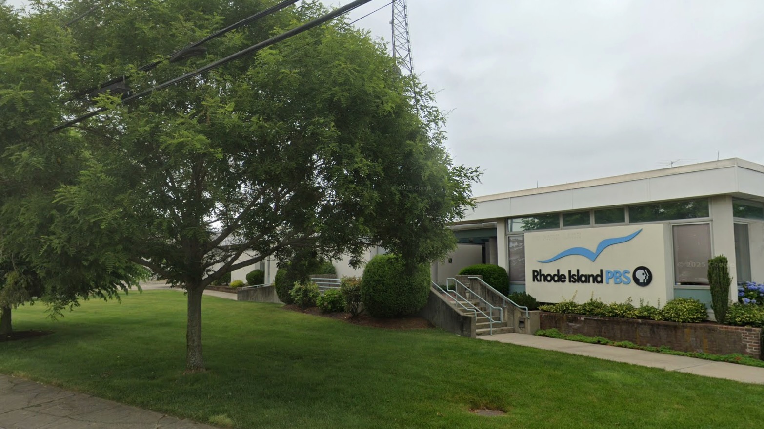 Single-story Rhode Island PBS building with a flat roof and large windows, surrounded by a well-kept lawn and trees. The station’s logo, featuring a blue wave and PBS emblem, is displayed on the front wall near the entrance.