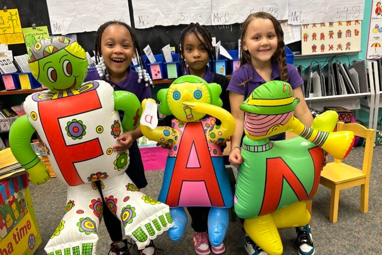 Three young children in a classroom smile and hold up large inflatable Letter People characters spelling “FAN.” The inflatables are brightly colored and designed with cartoon-style faces and outfits.