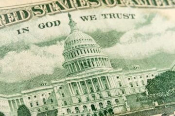 Close-up of the U.S. Capitol building as it appears on U.S. currency, showing detailed engraving with the words “In God We Trust” above.
