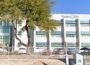 Exterior view of the Vegas PBS headquarters, a white multi-story building with large windows, trees in front, and cars parked along the fence line.