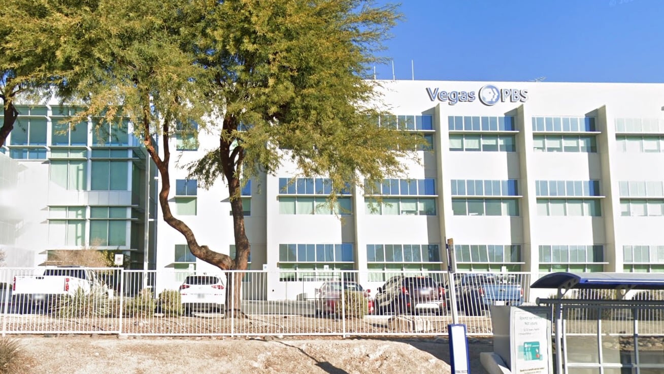 Exterior view of the Vegas PBS headquarters, a white multi-story building with large windows, trees in front, and cars parked along the fence line.