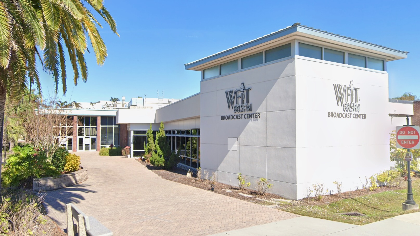 Exterior view of the WFIT 89.5 FM Broadcast Center, a modern low-rise building with large windows, surrounded by palm trees and landscaped greenery under a clear blue sky.