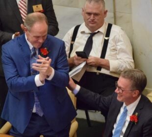In a legislative chamber, Rep. Johnny Rye, wearing a blue suit with a red carnation pinned to his lapel, claps and leans toward Rep. Carlton Wing, seated beside him in a dark suit with glasses and a blue tie. Wing smiles and places a hand on Rye’s arm. Other lawmakers and staff, including a man in suspenders using a phone, are visible in the background.