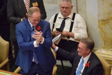 In a legislative chamber, Rep. Johnny Rye, wearing a blue suit with a red carnation pinned to his lapel, claps and leans toward Rep. Carlton Wing, seated beside him in a dark suit with glasses and a blue tie. Wing smiles and places a hand on Rye’s arm. Other lawmakers and staff, including a man in suspenders using a phone, are visible in the background.