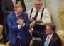 In a legislative chamber, Rep. Johnny Rye, wearing a blue suit with a red carnation pinned to his lapel, claps and leans toward Rep. Carlton Wing, seated beside him in a dark suit with glasses and a blue tie. Wing smiles and places a hand on Rye’s arm. Other lawmakers and staff, including a man in suspenders using a phone, are visible in the background.
