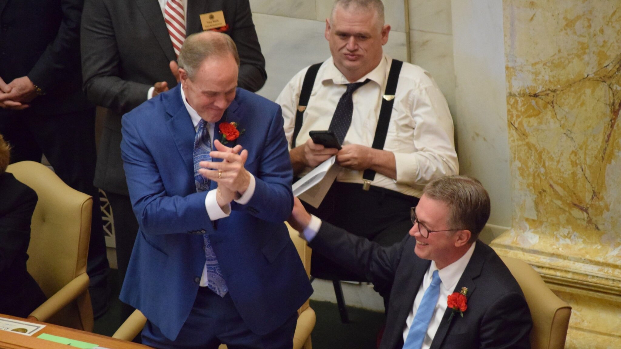 In a legislative chamber, Rep. Johnny Rye, wearing a blue suit with a red carnation pinned to his lapel, claps and leans toward Rep. Carlton Wing, seated beside him in a dark suit with glasses and a blue tie. Wing smiles and places a hand on Rye’s arm. Other lawmakers and staff, including a man in suspenders using a phone, are visible in the background.