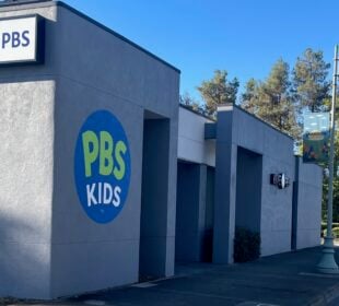 Exterior view of KIXE PBS headquarters in Redding, California. The gray building features a large circular PBS Kids logo painted on the wall and a mounted KIXE PBS sign. A tree-lined street with lampposts and banners runs alongside the building under a clear blue sky.