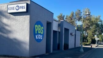 Exterior view of KIXE PBS headquarters in Redding, California. The gray building features a large circular PBS Kids logo painted on the wall and a mounted KIXE PBS sign. A tree-lined street with lampposts and banners runs alongside the building under a clear blue sky.