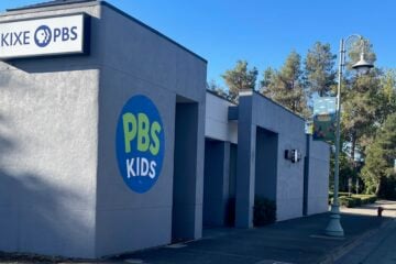 Exterior view of KIXE PBS headquarters in Redding, California. The gray building features a large circular PBS Kids logo painted on the wall and a mounted KIXE PBS sign. A tree-lined street with lampposts and banners runs alongside the building under a clear blue sky.