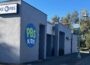 Exterior view of KIXE PBS headquarters in Redding, California. The gray building features a large circular PBS Kids logo painted on the wall and a mounted KIXE PBS sign. A tree-lined street with lampposts and banners runs alongside the building under a clear blue sky.