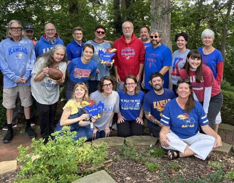 A group of Kansas Public Radio staff members pose outdoors during the Radio Rivals campaign in Lawrence, Kansas. They are wearing University of Kansas shirts in blue, red, and gray, with some holding props including a football and Jayhawk cutouts. The group smiles and kneels or stands together in front of trees and greenery, showing team spirit.