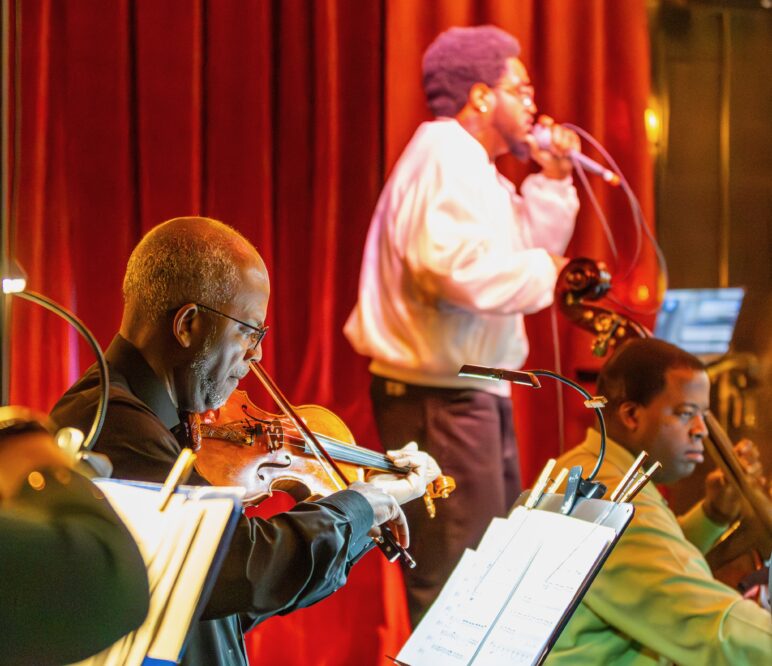 A violinist and cellist from the Charlotte Strings Collective perform onstage at Free Range Brewing while a vocalist sings into a microphone, with sheet music lit in front of the musicians and red stage curtains in the background.