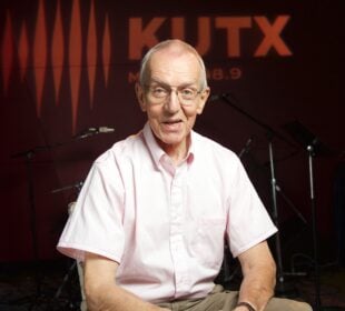 Wearing glasses and a light pink short-sleeve button-down shirt, John Aielli sits and smiles in a radio studio. Behind him, the wall is lit with the orange KUTX logo and sound wave graphic, with microphones, stands, and amplifiers visible in the background.