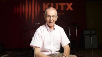 Wearing glasses and a light pink short-sleeve button-down shirt, John Aielli sits and smiles in a radio studio. Behind him, the wall is lit with the orange KUTX logo and sound wave graphic, with microphones, stands, and amplifiers visible in the background.