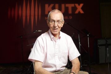 Wearing glasses and a light pink short-sleeve button-down shirt, John Aielli sits and smiles in a radio studio. Behind him, the wall is lit with the orange KUTX logo and sound wave graphic, with microphones, stands, and amplifiers visible in the background.