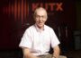 Wearing glasses and a light pink short-sleeve button-down shirt, John Aielli sits and smiles in a radio studio. Behind him, the wall is lit with the orange KUTX logo and sound wave graphic, with microphones, stands, and amplifiers visible in the background.