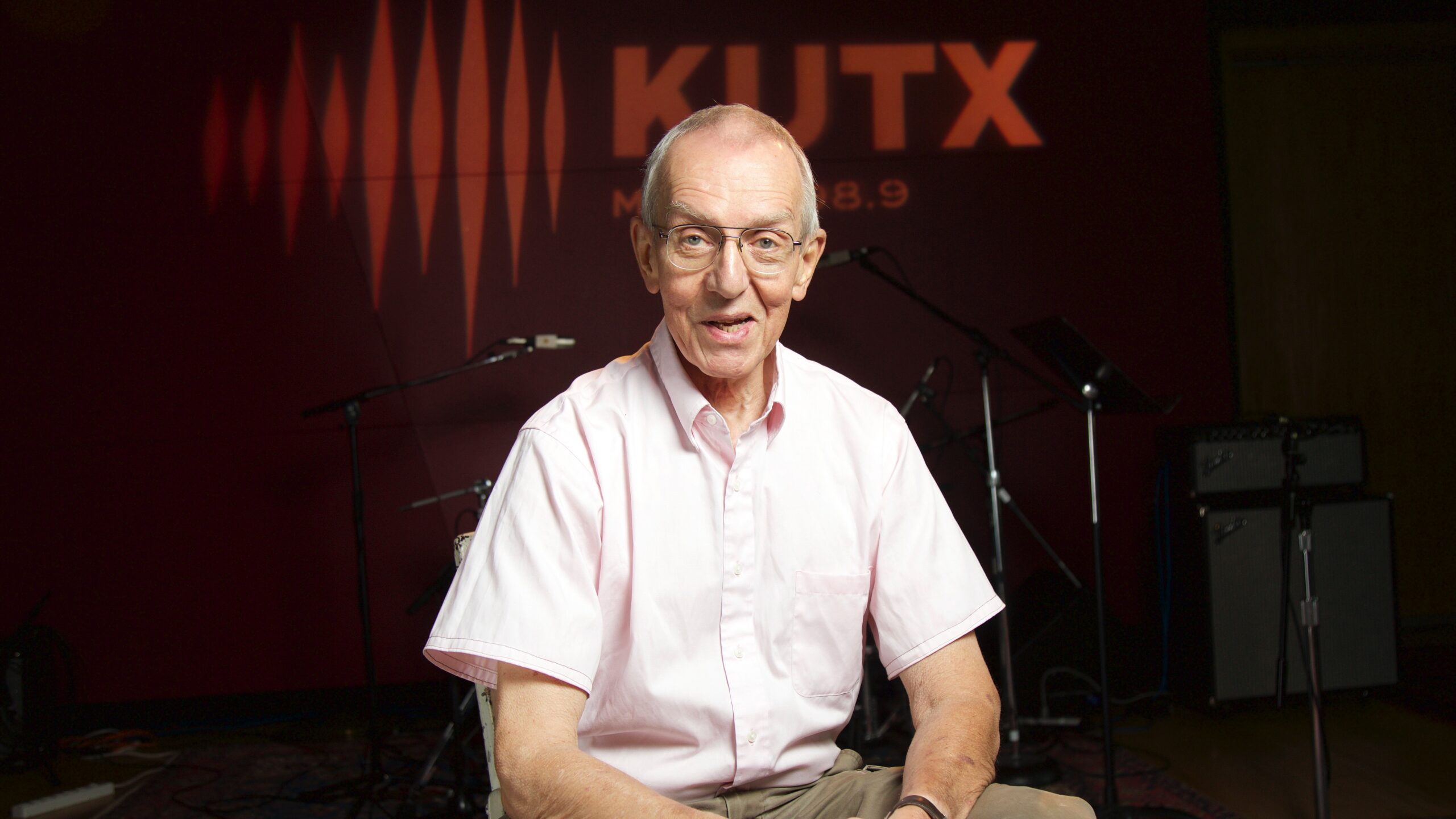 Wearing glasses and a light pink short-sleeve button-down shirt, John Aielli sits and smiles in a radio studio. Behind him, the wall is lit with the orange KUTX logo and sound wave graphic, with microphones, stands, and amplifiers visible in the background.