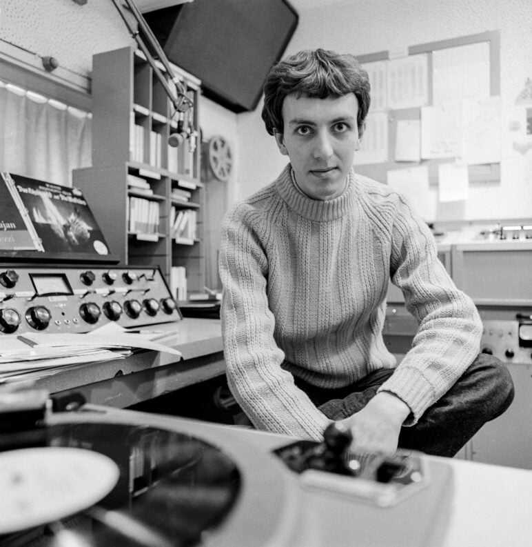 Wearing a cable-knit sweater, John Aielli sits in a radio control room. He looks directly at the camera, surrounded by equipment including a turntable in the foreground, a control panel with dials, and shelves of records and papers in the background.