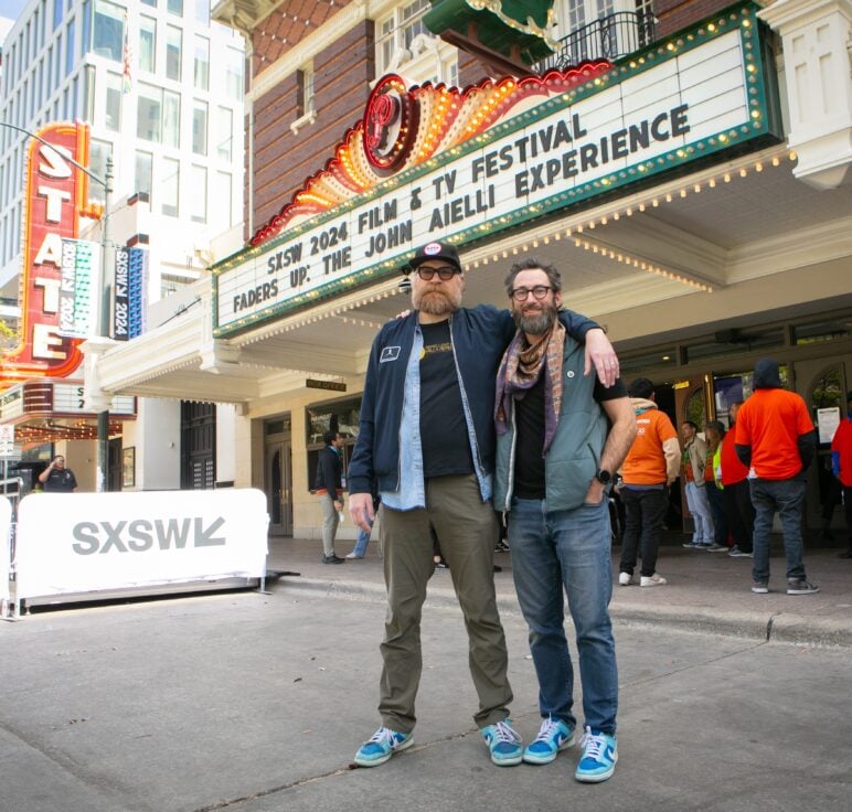 Sam Wainwright Douglas and David Hartstein stand on the sidewalk outside the Paramount Theatre in Austin, Texas, during SXSW 2024. The marquee above them reads “SXSW 2024 Film & TV Festival Faders Up: The John Aielli Experience.” People in orange shirts and festival staff are visible in the background near the entrance.