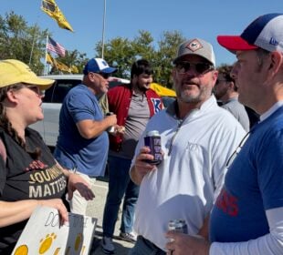 KBIA health reporter Rebecca Smith, wearing a yellow cap and black shirt with the words “Local journalism matters,” talks with two Kansas University football fans outside Memorial Stadium in Columbia, Missouri. One fan wears a white KU pullover and sunglasses while holding a drink, and another wears a blue KU shirt and white cap. Other fans and tailgating tents are visible in the background.