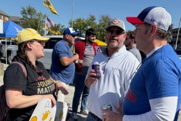 KBIA health reporter Rebecca Smith, wearing a yellow cap and black shirt with the words “Local journalism matters,” talks with two Kansas University football fans outside Memorial Stadium in Columbia, Missouri. One fan wears a white KU pullover and sunglasses while holding a drink, and another wears a blue KU shirt and white cap. Other fans and tailgating tents are visible in the background.