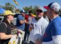 KBIA health reporter Rebecca Smith, wearing a yellow cap and black shirt with the words “Local journalism matters,” talks with two Kansas University football fans outside Memorial Stadium in Columbia, Missouri. One fan wears a white KU pullover and sunglasses while holding a drink, and another wears a blue KU shirt and white cap. Other fans and tailgating tents are visible in the background.