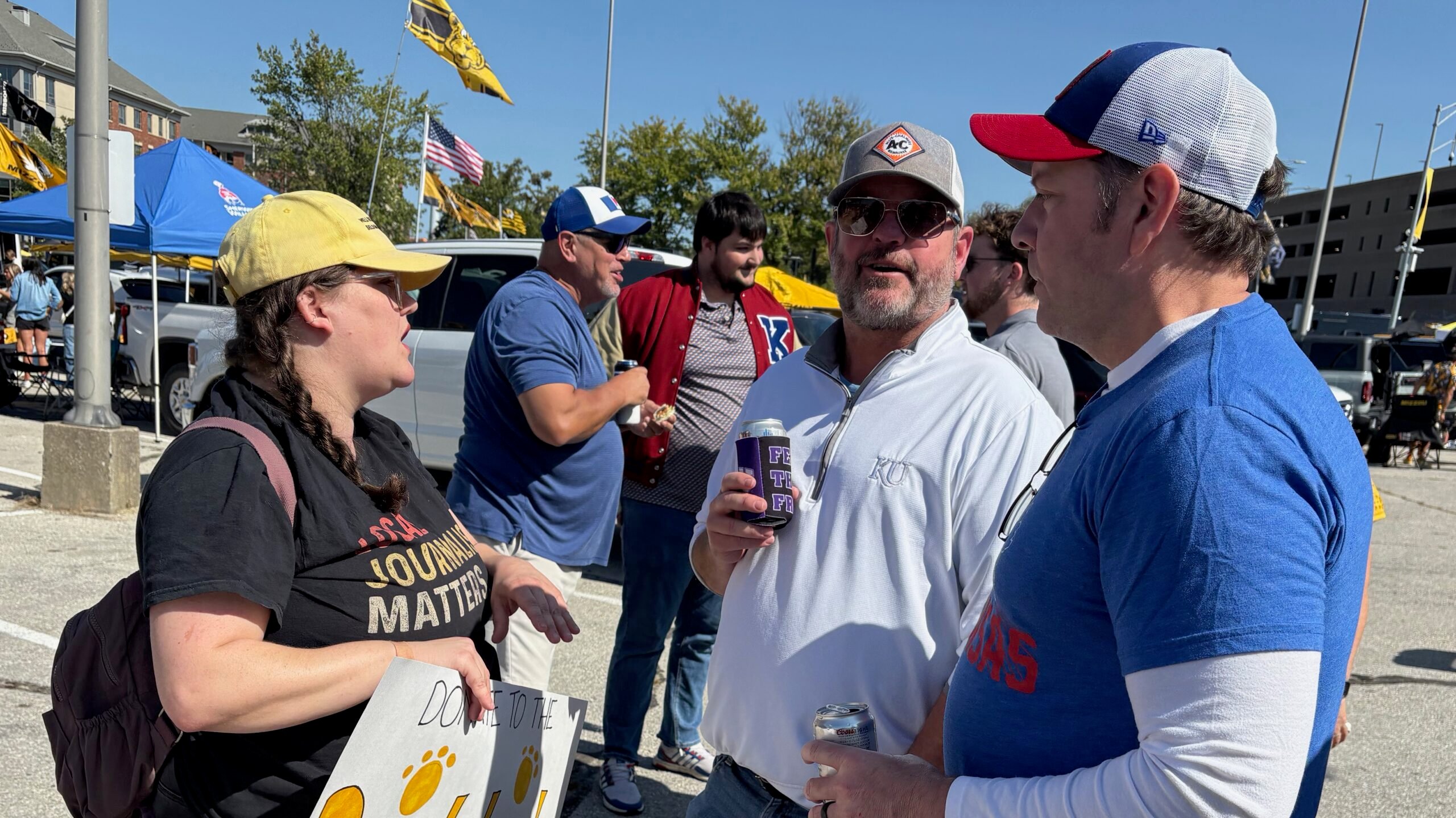 KBIA health reporter Rebecca Smith, wearing a yellow cap and black shirt with the words “Local journalism matters,” talks with two Kansas University football fans outside Memorial Stadium in Columbia, Missouri. One fan wears a white KU pullover and sunglasses while holding a drink, and another wears a blue KU shirt and white cap. Other fans and tailgating tents are visible in the background.