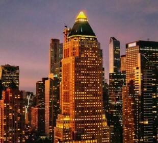 A cityscape at dusk shows the illuminated skyline of Manhattan. At the center stands One Worldwide Plaza, a tall building with a pyramid-shaped green roof topped by a glowing triangular light. The tower is surrounded by other high-rise buildings, their windows lit against the purple and orange evening sky.