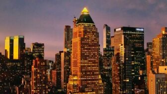 A cityscape at dusk shows the illuminated skyline of Manhattan. At the center stands One Worldwide Plaza, a tall building with a pyramid-shaped green roof topped by a glowing triangular light. The tower is surrounded by other high-rise buildings, their windows lit against the purple and orange evening sky.