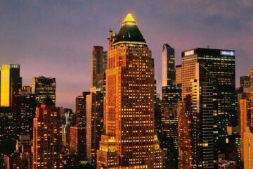 A cityscape at dusk shows the illuminated skyline of Manhattan. At the center stands One Worldwide Plaza, a tall building with a pyramid-shaped green roof topped by a glowing triangular light. The tower is surrounded by other high-rise buildings, their windows lit against the purple and orange evening sky.